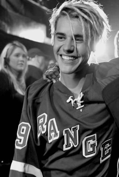Justin Bieber - preto e branco Foto preta e branca, Justin Bieber está sorrindo, vestindo uma camiseta de time, está sorrindo e abraçado com alguém.